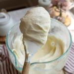 Close-up of fluffy homemade cream cheese frosting on a spatula, with a glass mixing bowl of frosting in the background—perfect for cakes, cupcakes, and desserts.