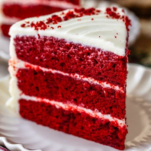 Slice of red velvet cake with cream cheese frosting on a white scalloped plate, showing vibrant red layers and smooth white icingโclassic Southern dessert perfect for holidays and special occasions.