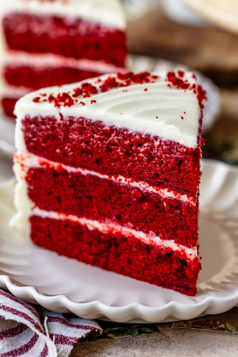 Slice of red velvet cake with cream cheese frosting on a white scalloped plate, showing vibrant red layers and smooth white icing—classic Southern dessert perfect for holidays and special occasions.