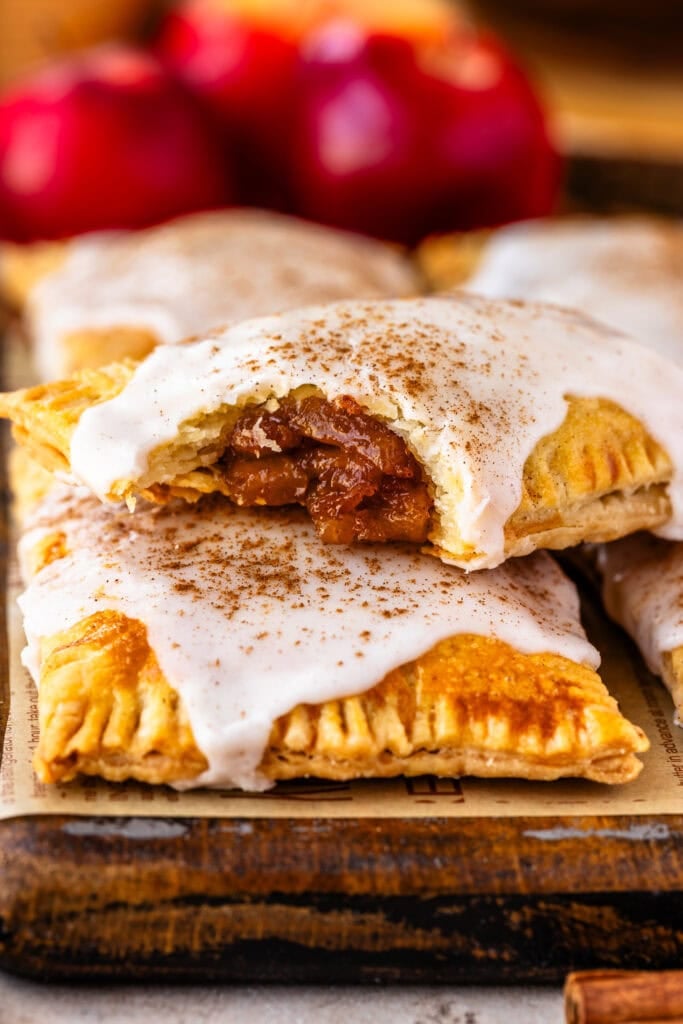 Golden brown apple pop tarts topped with white glaze and sprinkled with cinnamon, with one pastry showing the spiced apple filling inside.