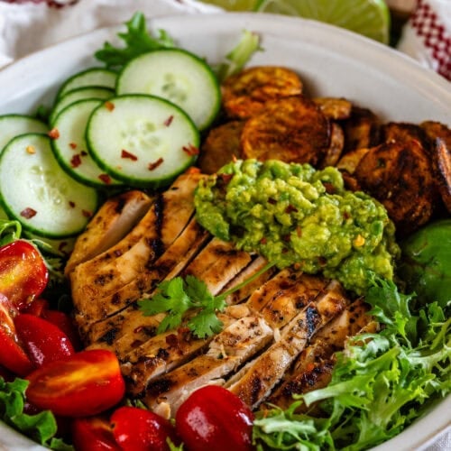 A bowl filled with grilled marinated chicken, fresh greens, cherry tomatoes, cucumber slices, guacamole, and crispy roasted potatoes, garnished with cilantro and lime.