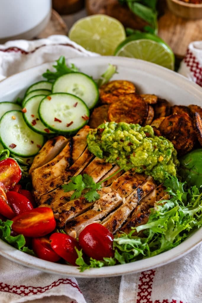 A bowl filled with grilled marinated chicken, fresh greens, cherry tomatoes, cucumber slices, guacamole, and crispy roasted potatoes, garnished with cilantro and lime.