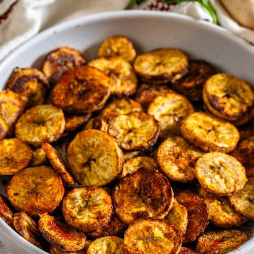 A white bowl filled with crispy baked plantain chips, golden brown and seasoned with spices.