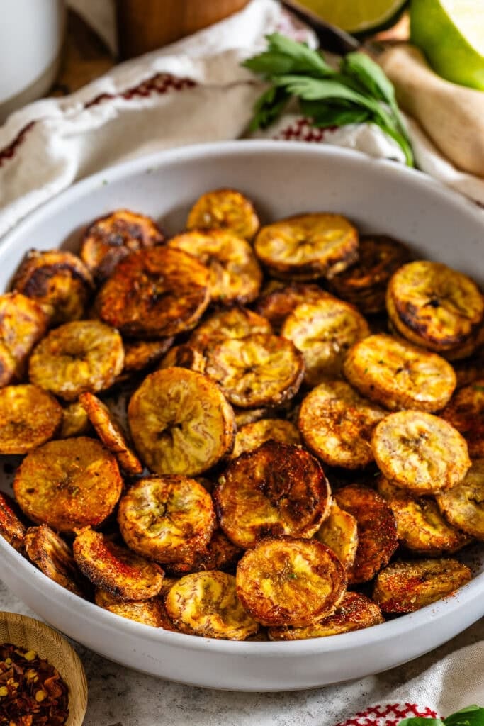 A white bowl filled with crispy baked plantain chips, golden brown and seasoned with spices.