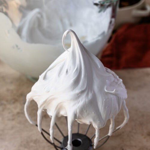 A whisk attachment covered in glossy marshmallow frosting with a tall peak, with a mixing bowl full of frosting in the background.