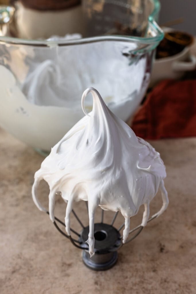 A whisk attachment covered in glossy marshmallow frosting with a tall peak, with a mixing bowl full of frosting in the background.