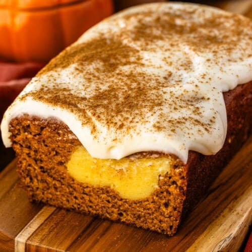 A loaf of pumpkin cream cheese bread on a wooden board, topped with cream cheese frosting and cinnamon, with a swirl of creamy cheesecake filling visible inside the bread.
