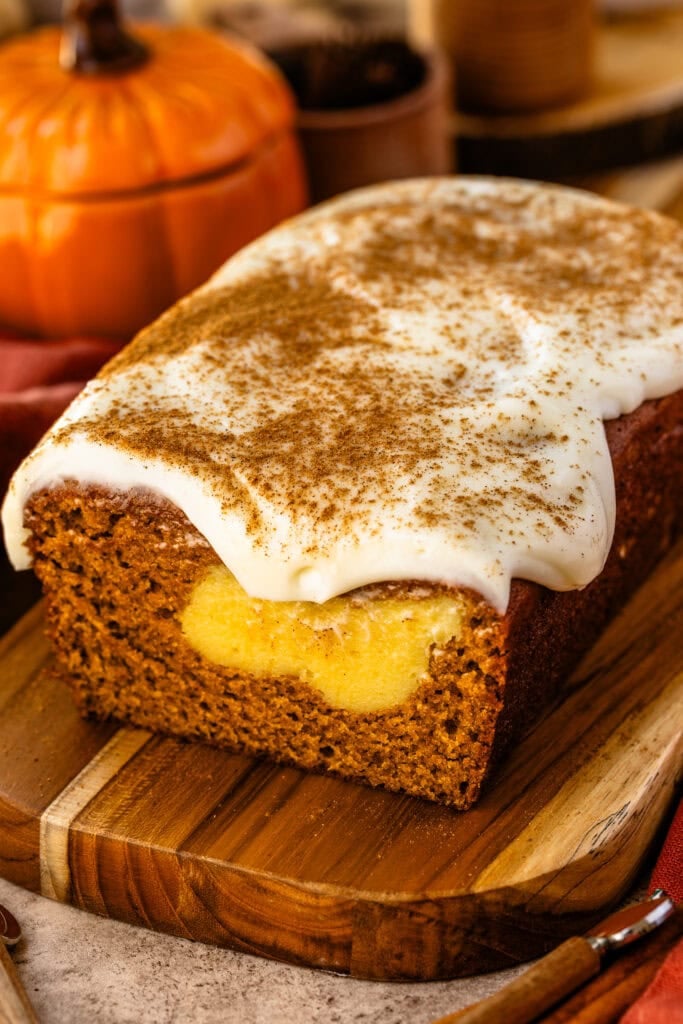A loaf of pumpkin cream cheese bread on a wooden board, topped with cream cheese frosting and cinnamon, with a swirl of creamy cheesecake filling visible inside the bread.