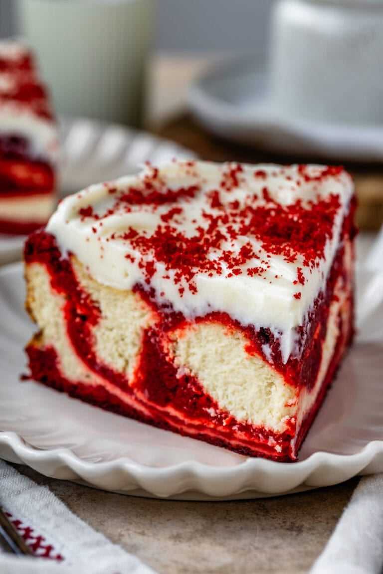 A slice of red velvet cheesecake on a white plate, topped with cream cheese frosting and sprinkled with red velvet crumbs.