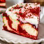 A slice of red velvet cheesecake on a white plate, topped with cream cheese frosting and sprinkled with red velvet crumbs.