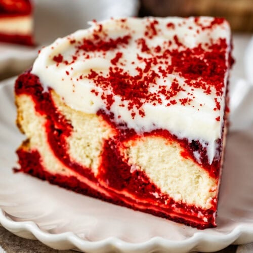 A slice of red velvet cheesecake on a white plate, topped with cream cheese frosting and sprinkled with red velvet crumbs.