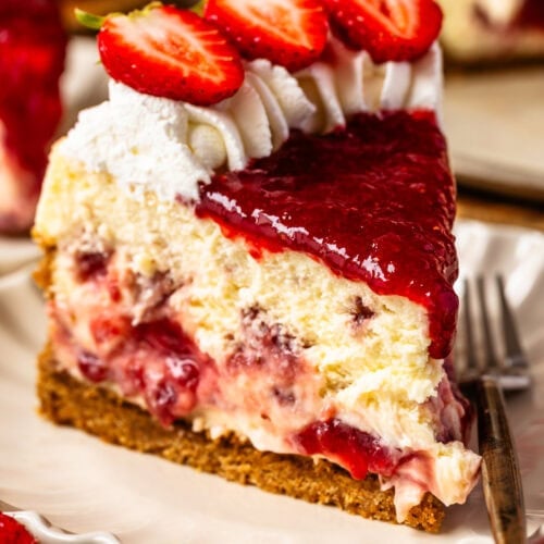 A slice of strawberry cheesecake on a plate, topped with whipped cream, fresh strawberry slices, and strawberry sauce, showing swirls of strawberry filling inside the creamy cheesecake.