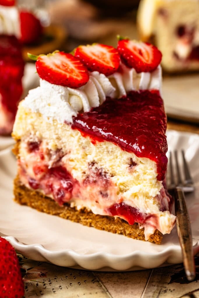 A slice of strawberry cheesecake on a plate, topped with whipped cream, fresh strawberry slices, and strawberry sauce, showing swirls of strawberry filling inside the creamy cheesecake.