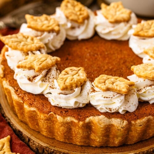 A baked sweet potato pie topped with swirls of whipped cream and decorative leaf-shaped pie crust cutouts, set on a wooden slice.