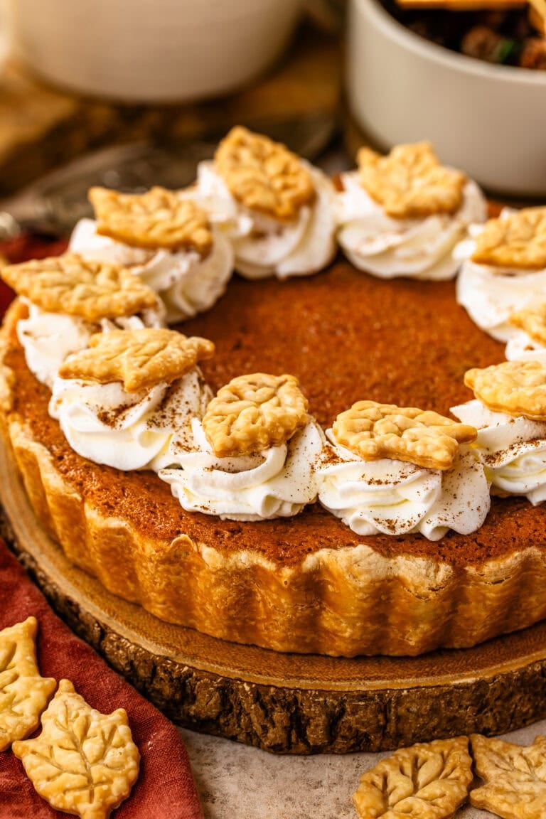 A baked sweet potato pie topped with swirls of whipped cream and decorative leaf-shaped pie crust cutouts, set on a wooden slice.