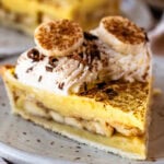 A close-up slice of banana cream pie on a ceramic plate, showing layers of bananas, vanilla custard, and whipped cream swirls topped with banana slices and chocolate shavings.