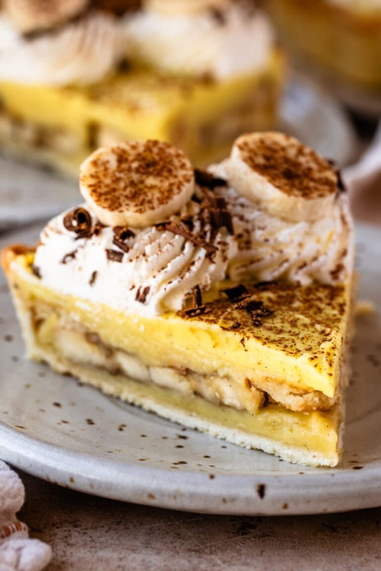 A close-up slice of banana cream pie on a ceramic plate, showing layers of bananas, vanilla custard, and whipped cream swirls topped with banana slices and chocolate shavings.