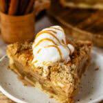 A slice of Dutch apple pie topped with a scoop of vanilla ice cream and drizzled with caramel sauce, served on a white plate with a cinnamon stick holder in the background.