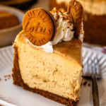A close-up slice of no-bake Biscoff cheesecake topped with whipped cream, Biscoff cookies, and cookie crumbs, showing its creamy texture and crumbly cookie crust.