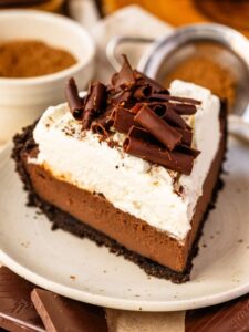 A close-up of a slice of chocolate cream pie on a plate. The pie features a crisp Oreo cookie crust, a silky smooth chocolate custard filling, a thick layer of fluffy whipped cream, and a generous topping of dark chocolate curls for an elegant finish.