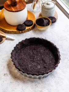 A close-up of a tart pan filled with a pressed Oreo cookie crust on a light countertop, surrounded by a bowl of Oreos and rustic kitchen decor.