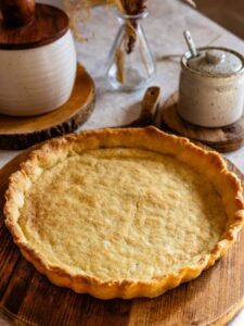 A close-up of a golden baked pie crust resting on a wooden board, showing its flaky texture and crimped edges.