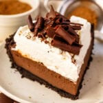 A close-up of a slice of chocolate cream pie on a plate. The pie features a crisp Oreo cookie crust, a silky smooth chocolate custard filling, a thick layer of fluffy whipped cream, and a generous topping of dark chocolate curls for an elegant finish.