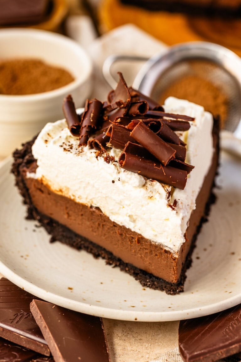 A close-up of a slice of chocolate cream pie on a plate. The pie features a crisp Oreo cookie crust, a silky smooth chocolate custard filling, a thick layer of fluffy whipped cream, and a generous topping of dark chocolate curls for an elegant finish.