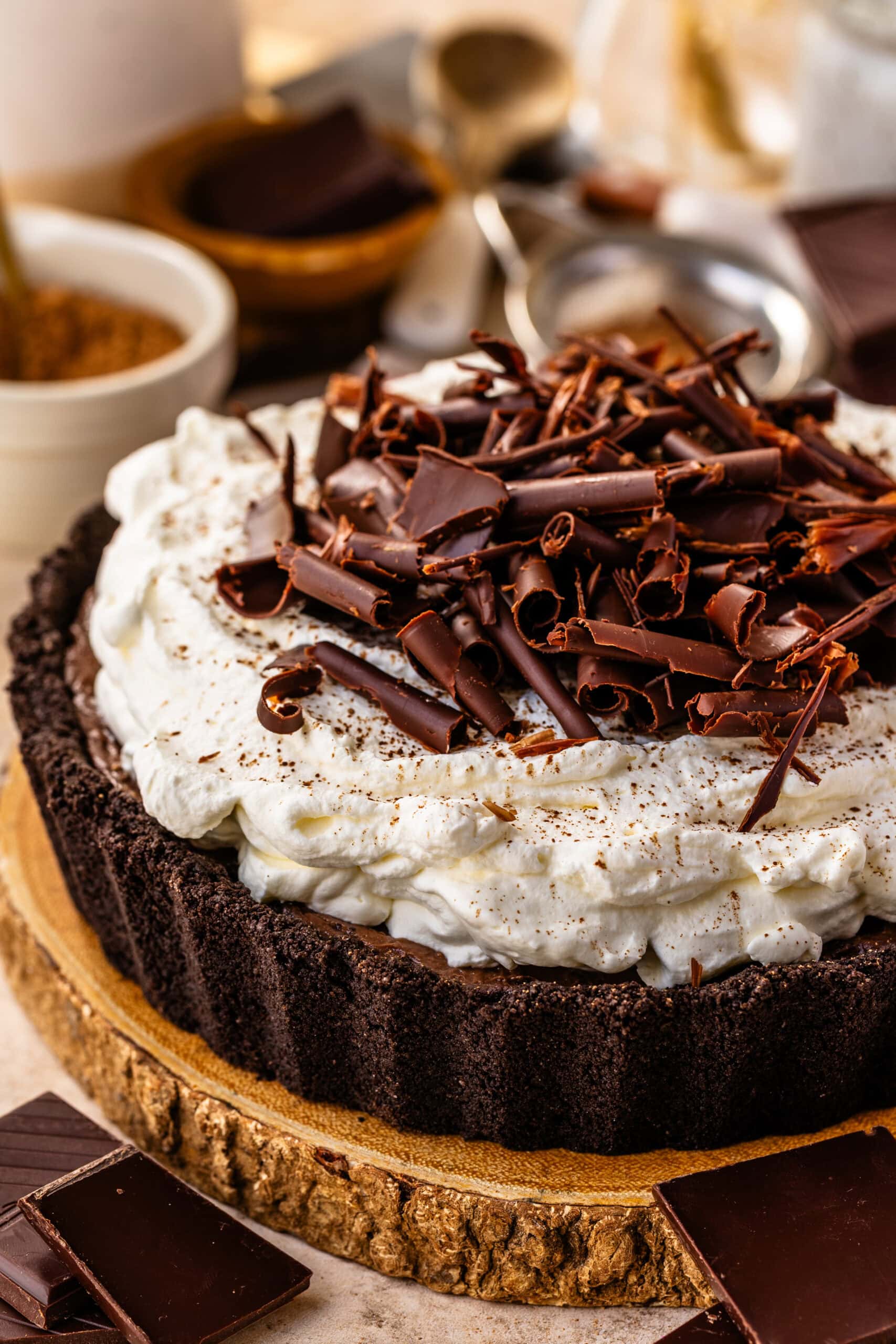 Close-up of a whole chocolate cream pie on a wooden board. The pie features a dark Oreo crust filled with silky chocolate custard, topped with a thick layer of fluffy whipped cream and an elegant pile of dark chocolate curls for a beautiful, decadent finish.