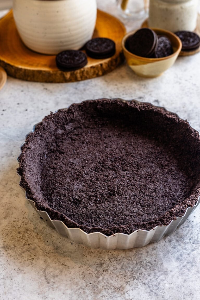 A close-up of a tart pan filled with a pressed Oreo cookie crust on a light countertop, surrounded by a bowl of Oreos and rustic kitchen decor.
