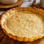 A close-up of a golden baked pie crust resting on a wooden board, showing its flaky texture and crimped edges.