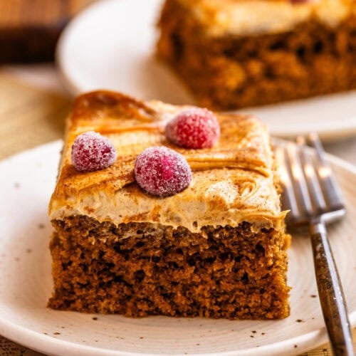 A slice of gingerbread cake topped with creamy gingerbread frosting, caramel swirls, and sugared cranberries on a plate.