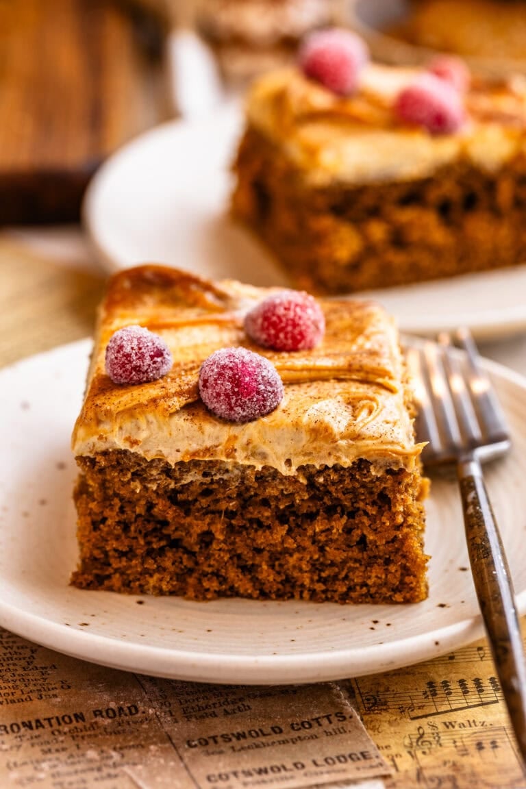 A slice of gingerbread cake topped with creamy gingerbread frosting, caramel swirls, and sugared cranberries on a plate.