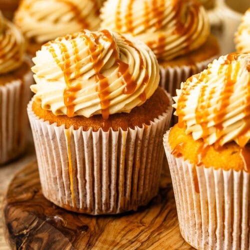 A close-up of frosted salted caramel cupcakes topped with swirls of caramel buttercream and drizzled with golden caramel sauce, arranged together on a rustic surface.
