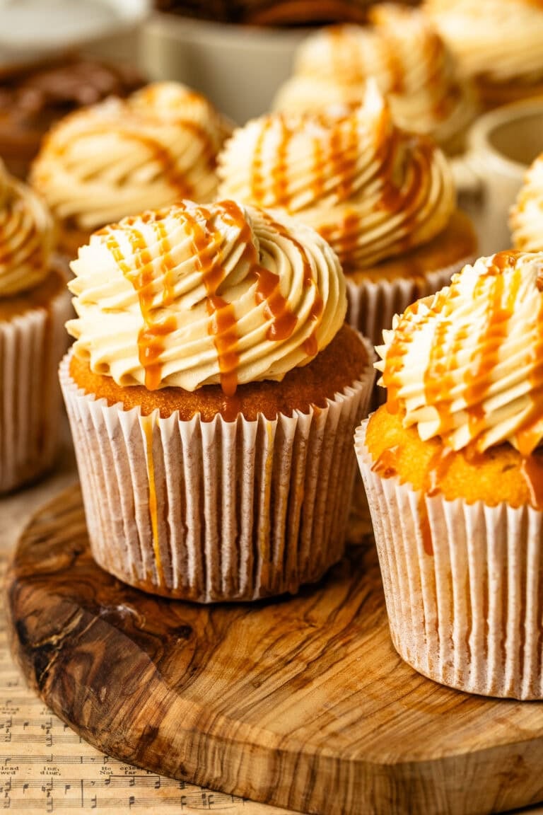 A close-up of frosted salted caramel cupcakes topped with swirls of caramel buttercream and drizzled with golden caramel sauce, arranged together on a rustic surface.