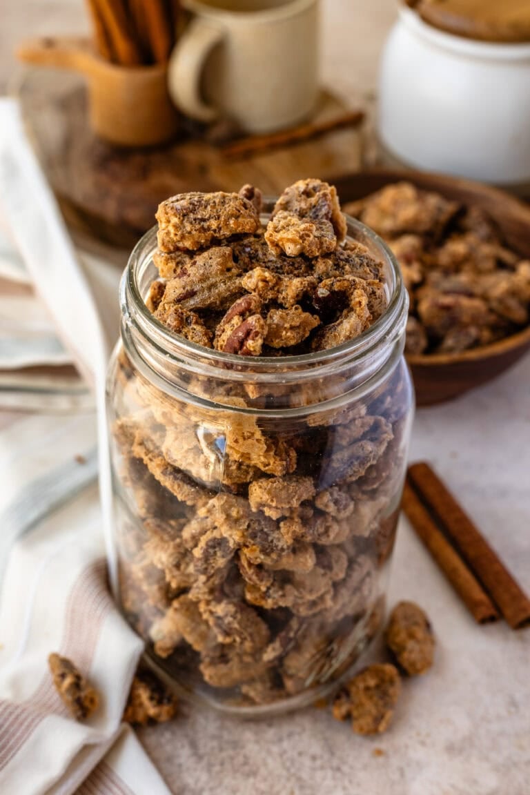 A glass jar filled to the top with crunchy candied pecans, with extra pecans and cinnamon sticks scattered around the table.