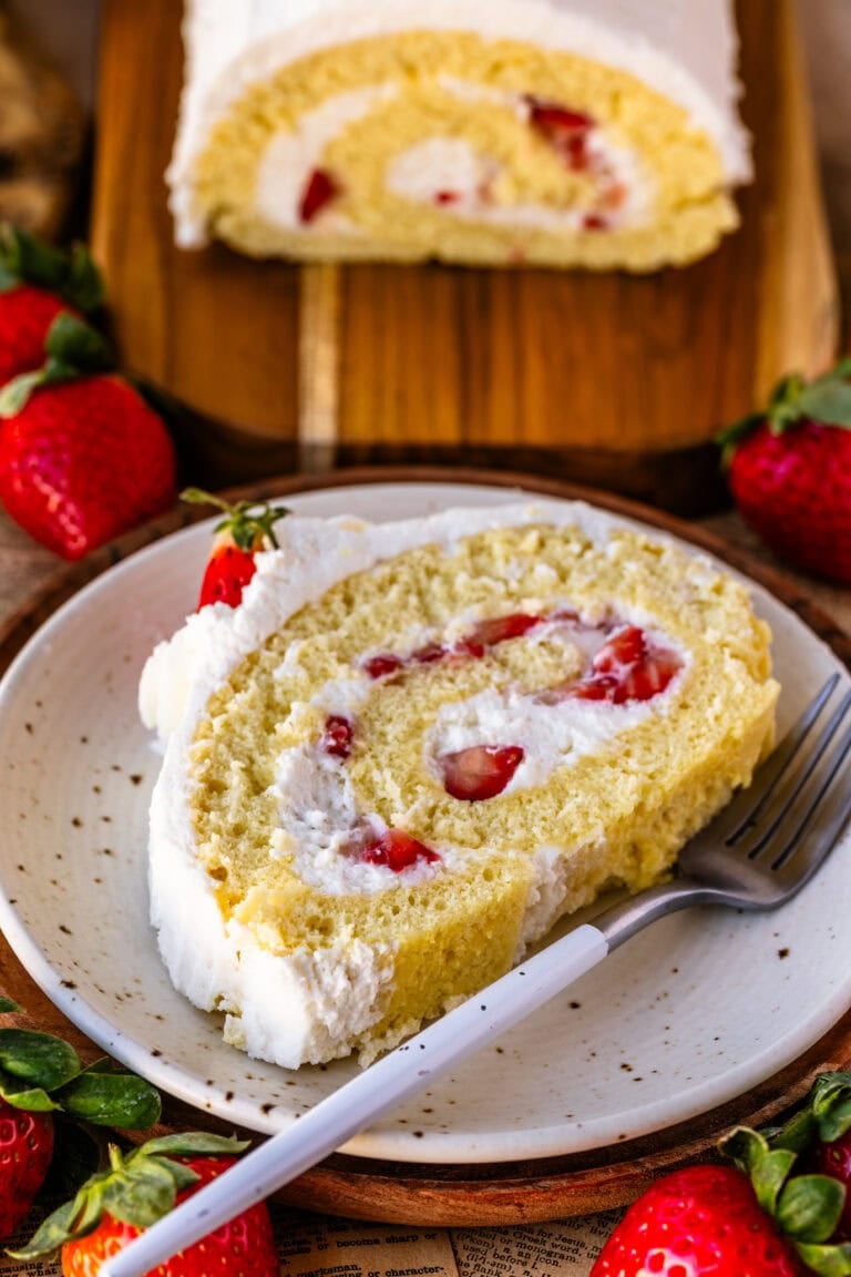 A slice of vanilla Swiss roll cake filled with whipped cream and fresh strawberries on a white plate.