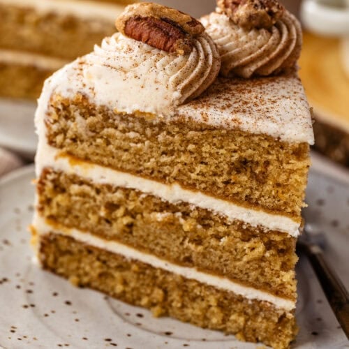 Slice of spiced layer cake with cream cheese frosting, topped with piped frosting and candied pecans.