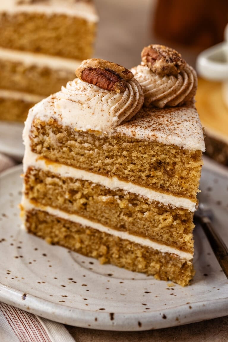 Slice of spiced layer cake with cream cheese frosting, topped with piped frosting and candied pecans.