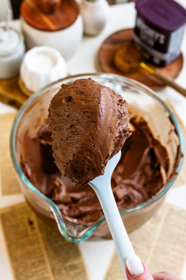 A spoonful of creamy chocolate buttercream frosting held above a glass bowl.