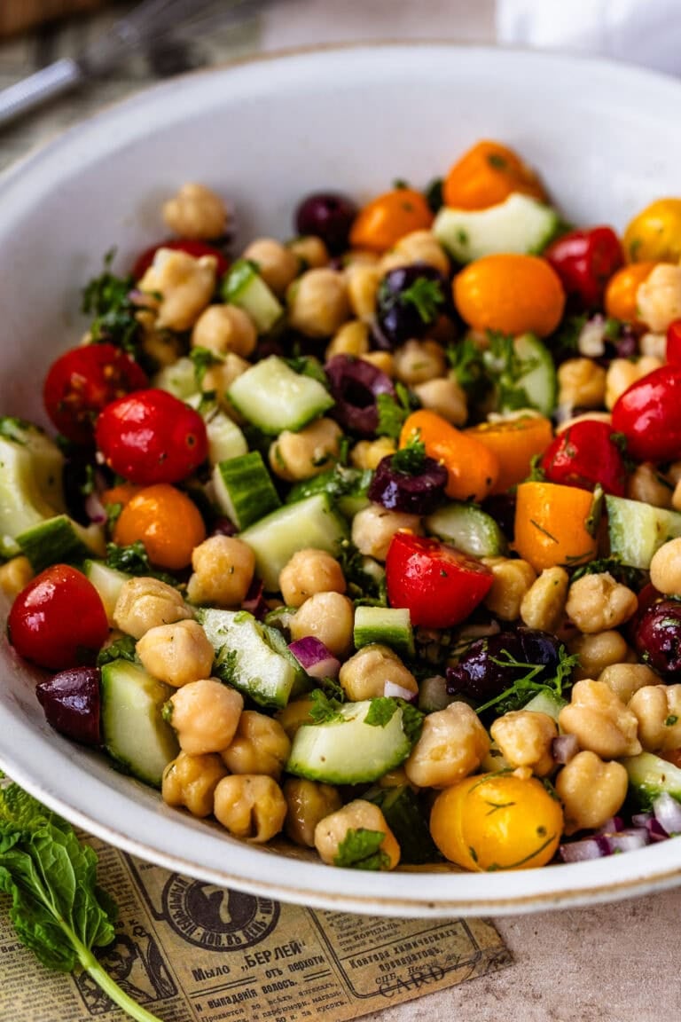 Chickpea salad with cucumbers, cherry tomatoes, olives, red onion, and fresh herbs in a white bowl.