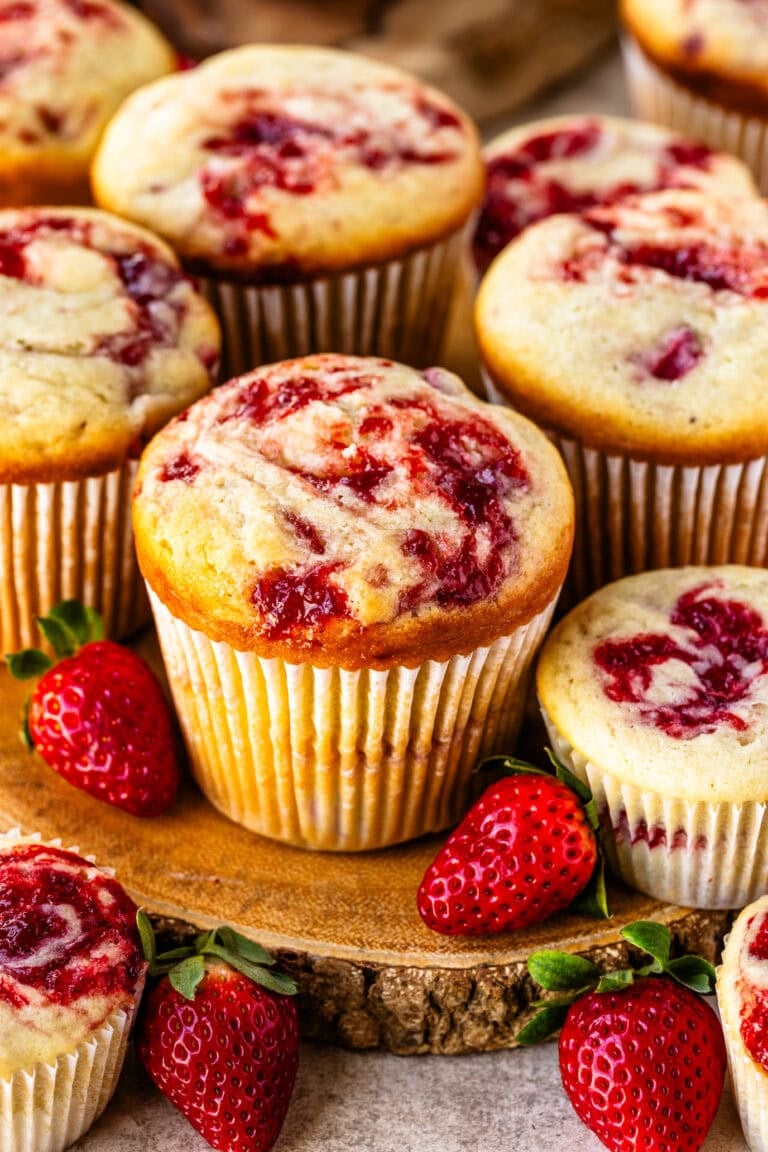 Strawberry cream cheese muffins with a golden top and visible strawberry swirls, arranged on a wooden board with fresh strawberries.