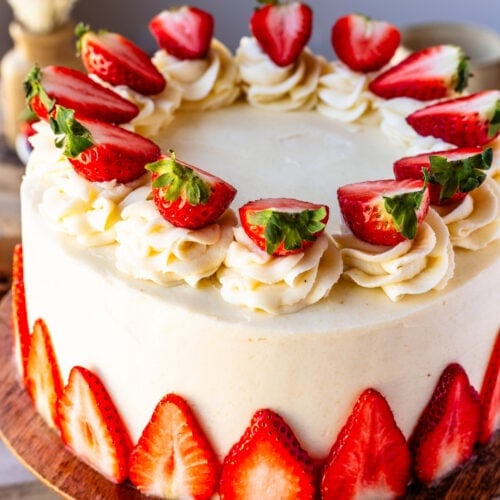 An entire Almond Strawberry Cake beautifully decorated with frosting and strawberries on top of a wooden cake mount.