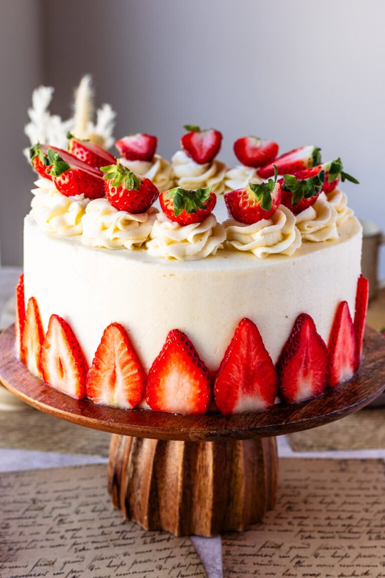 An entire Almond Strawberry Cake beautifully decorated with frosting and strawberries on top of a wooden cake mount.