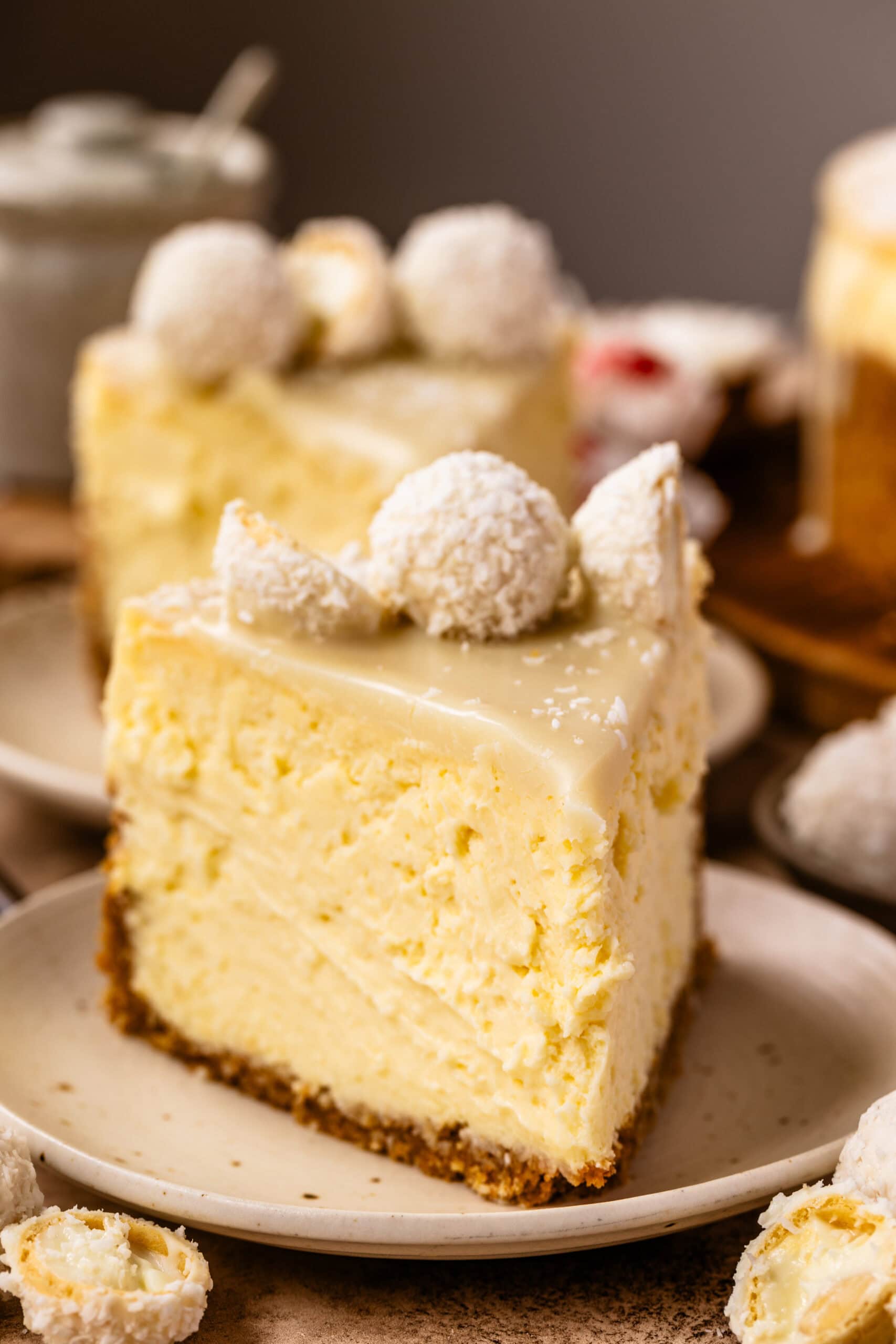 Slice of coconut cheesecake with white chocolate ganache and coconut truffles on top, served on a plate with coconut candies around it.