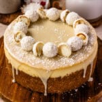 A whole coconut cheesecake with white chocolate ganache and coconut truffles on top, resting on a wooden serving board.