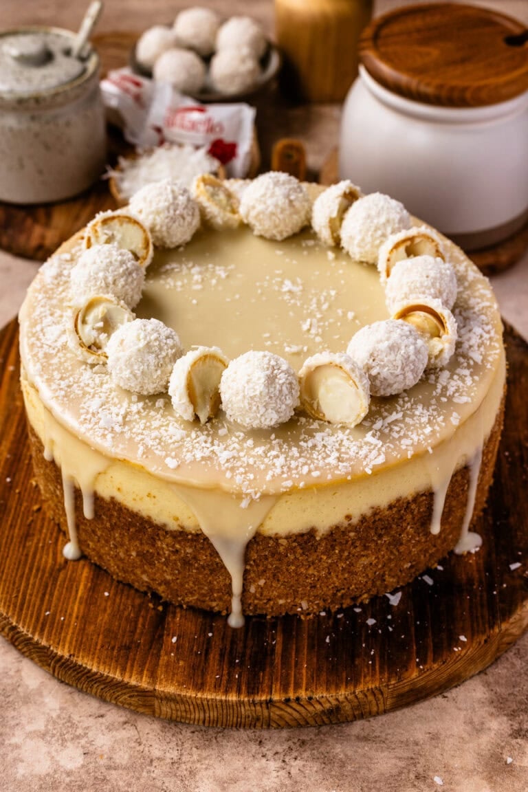 A whole coconut cheesecake with white chocolate ganache and coconut truffles on top, resting on a wooden serving board.