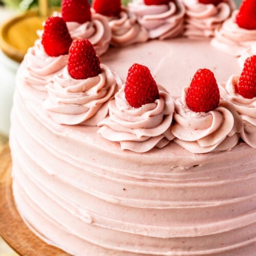 A full, delicately frosted Raspberry Cake topped with fresh strawberries on a wooden cake stand.