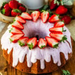 A whole strawberry bundt cake topped with strawberry cream cheese frosting and halved fresh strawberries arranged on top.
