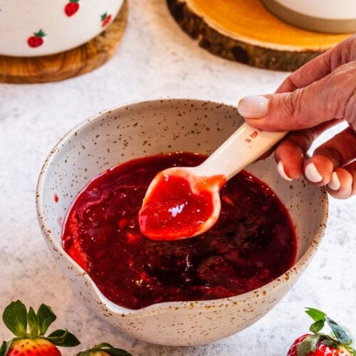 A spoon lifting thick homemade strawberry sauce from a bowl, with fresh strawberries in the background.
